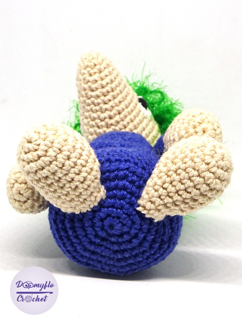 May include: A crocheted toy in the shape of a blue bird with beige wings and a green and white head. The bird is lying on its back with its wings spread out.