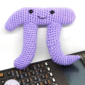 Peut inclure: Un jouet en peluche en crochet violet en forme de symbole mathématique pi. Le jouet a un visage souriant avec deux yeux noirs.