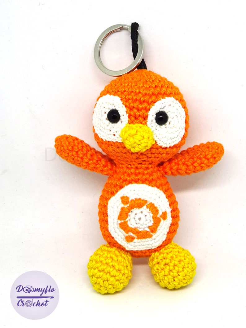 May include: A handmade orange crochet bird keychain with white accents and black eyes. The bird has yellow feet and a yellow beak. The keychain includes a silver metal ring and a black string. The bird's belly has a white circle with an orange design.