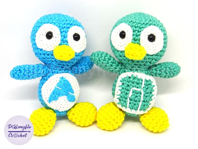 May include: Two handmade crochet penguin dolls. One is blue with a white belly and the letter "A", the other is green with a white belly and the letter "H". Both have yellow feet, white eyes, and yellow beaks. The dolls are handmade.