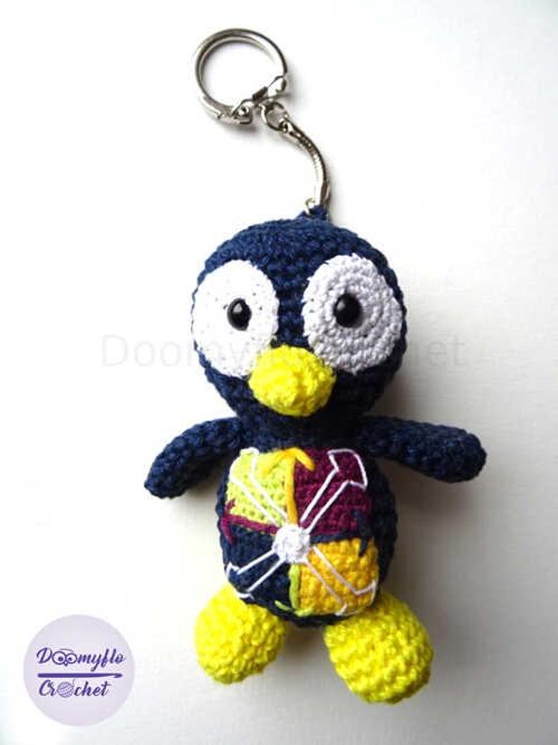May include: Handmade crochet penguin keychain. The penguin is navy blue with white eyes, a yellow beak, and yellow feet. The penguin's belly features a colorful design. A silver keyring is attached to the top of the penguin.