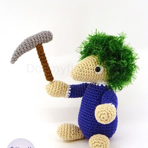 May include: A crocheted toy with green hair, blue overalls, and a gray pickaxe. The toy is sitting on a white surface.