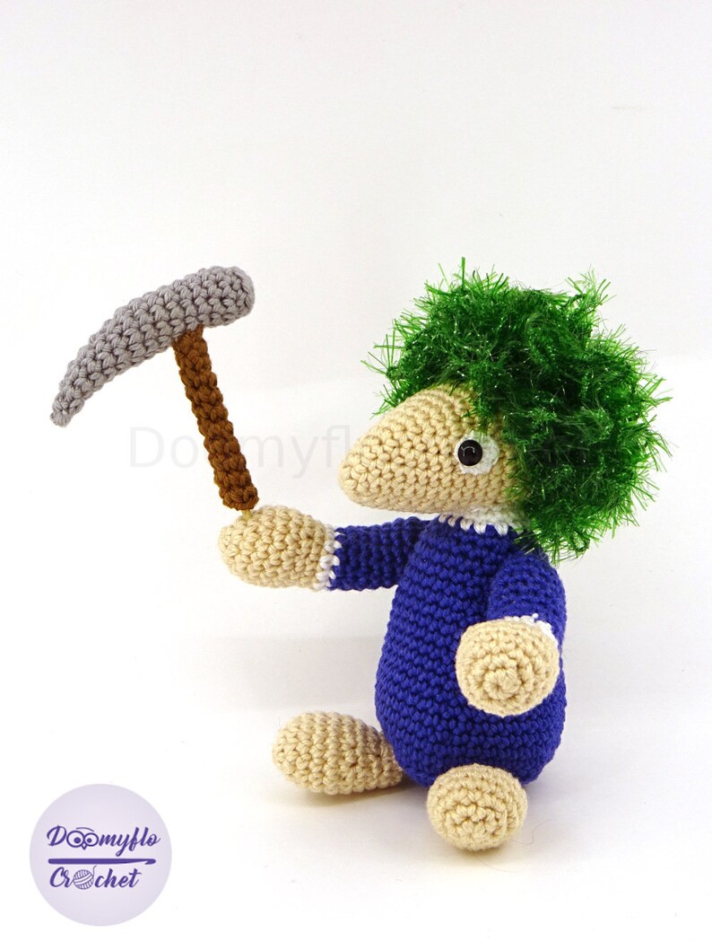 May include: A crocheted toy with green hair, blue overalls, and a gray pickaxe. The toy is sitting on a white surface.