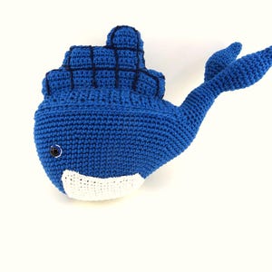 Peut inclure: Un jouet en peluche baleine bleu crocheté avec un ventre blanc et un œil noir. La baleine a une nageoire sur le dos et une queue.