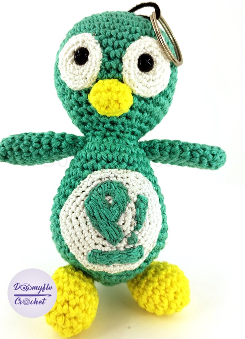 May include: Crocheted penguin keychain in teal, white, and yellow. The penguin has large white eyes with black pupils, a yellow beak, and yellow feet. The penguin's belly features a white circle with a teal number 9. The keychain has a metal ring.