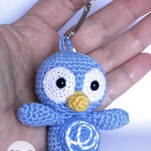 May include: A handmade blue crochet penguin keychain with white accents, black eyes, and a yellow beak and feet. The keychain has a silver clasp and a spring coil. The penguin has a white design on its belly. The logo "Doomyflo Crochet" is visible.