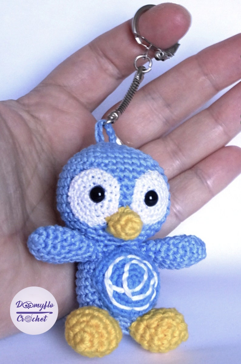 May include: A handmade blue crochet penguin keychain with white accents, black eyes, and a yellow beak and feet. The keychain has a silver clasp and a spring coil. The penguin has a white design on its belly. The logo "Doomyflo Crochet" is visible.