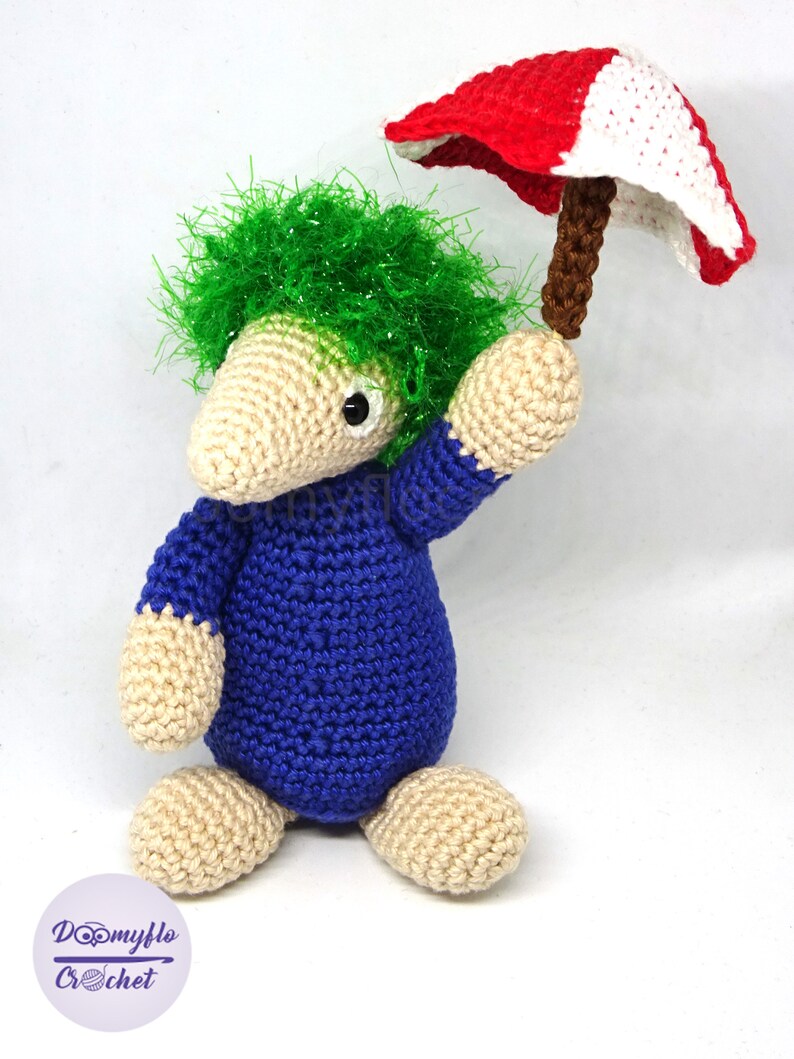 May include: A crocheted green-haired creature wearing a blue jumpsuit holds a red and white crocheted umbrella.