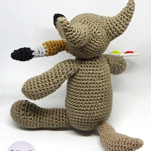 Puede incluir: Un lobo de crochet marr&oacute;n sosteniendo un pincel y una paleta con pintura. El lobo est&aacute; sentado sobre una superficie blanca.