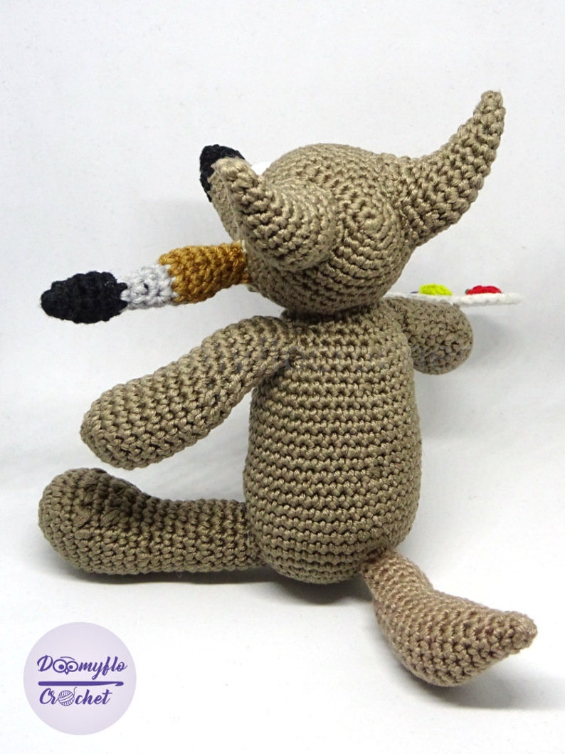 Puede incluir: Un lobo de crochet marr&oacute;n sosteniendo un pincel y una paleta con pintura. El lobo est&aacute; sentado sobre una superficie blanca.