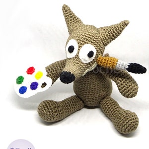 Puede incluir: Un lobo de crochet marr&oacute;n sosteniendo una paleta de pintura blanca con puntos de pintura de colores y un pincel. El lobo tiene ojos grandes blancos con pupilas negras.