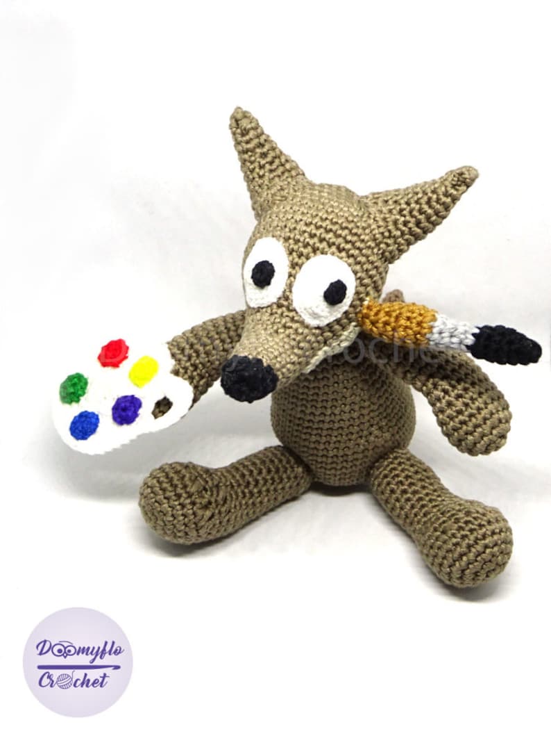 Puede incluir: Un lobo de crochet marr&oacute;n sosteniendo una paleta de pintura blanca con puntos de pintura de colores y un pincel. El lobo tiene ojos grandes blancos con pupilas negras.