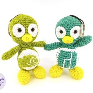 May include: Two handmade crochet penguin keychains. One is green with white accents, the other is teal with white accents. Both have yellow feet and beaks, black eyes, and a keychain ring. The keychains are standing on a white surface.