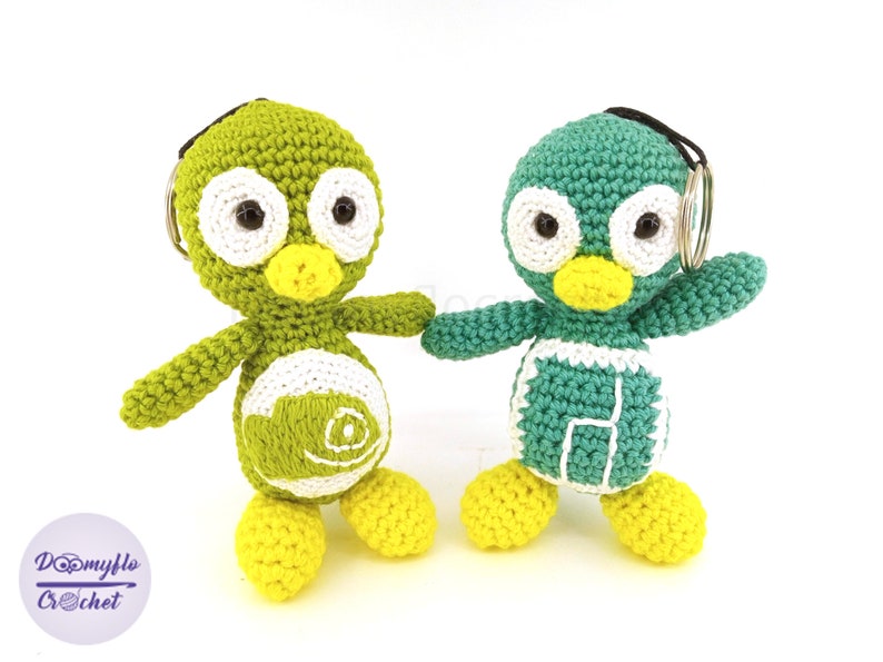 May include: Two handmade crochet penguin keychains. One is green with white accents, the other is teal with white accents. Both have yellow feet and beaks, black eyes, and a keychain ring. The keychains are standing on a white surface.