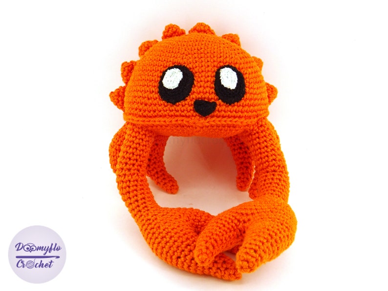May include: Orange crocheted crab with black eyes and a smiling mouth. The crab is sitting on a white background.