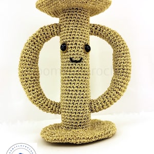 Peut inclure: Un jouet amigurumi en crochet doré en forme de lettre grecque Phi. Le jouet a un visage souriant avec des yeux noirs et une bouche noire.