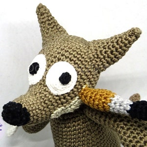 Puede incluir: Un lobo de peluche tejido a crochet con grandes ojos blancos y una nariz negra. El lobo es marr&oacute;n y tiene una cola larga y delgada con rayas negras, blancas y doradas.