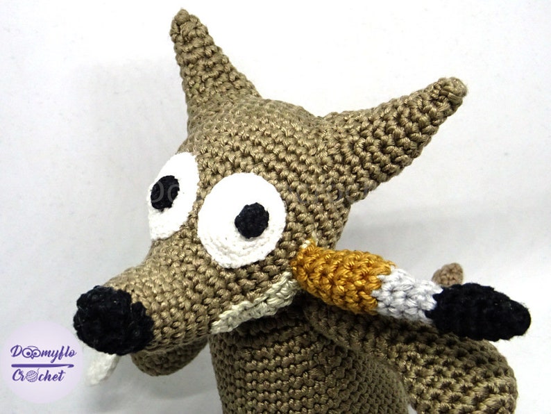 Puede incluir: Un lobo de peluche tejido a crochet con grandes ojos blancos y una nariz negra. El lobo es marr&oacute;n y tiene una cola larga y delgada con rayas negras, blancas y doradas.