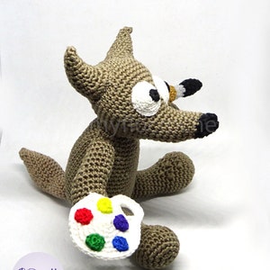 Puede incluir: Un lobo marr&oacute;n de crochet sosteniendo una paleta de pintura blanca con pintura roja, amarilla, verde, azul y morada. El lobo tiene ojos grandes blancos y una nariz negra.