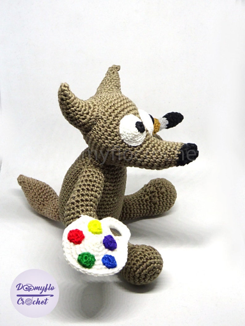 Puede incluir: Un lobo marr&oacute;n de crochet sosteniendo una paleta de pintura blanca con pintura roja, amarilla, verde, azul y morada. El lobo tiene ojos grandes blancos y una nariz negra.