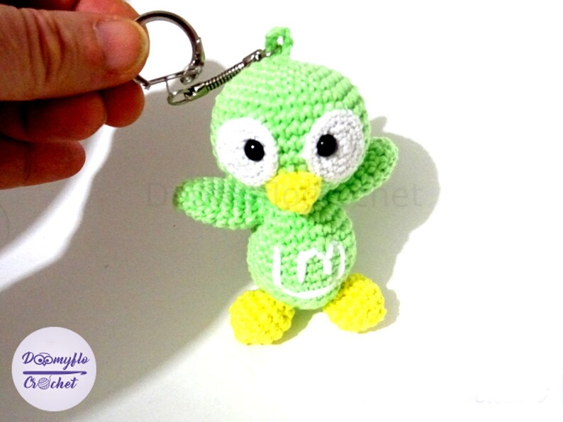 May include: A handmade green crochet bird keychain with white accents and black eyes. The bird has yellow feet and a yellow beak. The keychain is attached to a silver metal ring. The bird is holding its arms out. The keychain is a cute accessory.