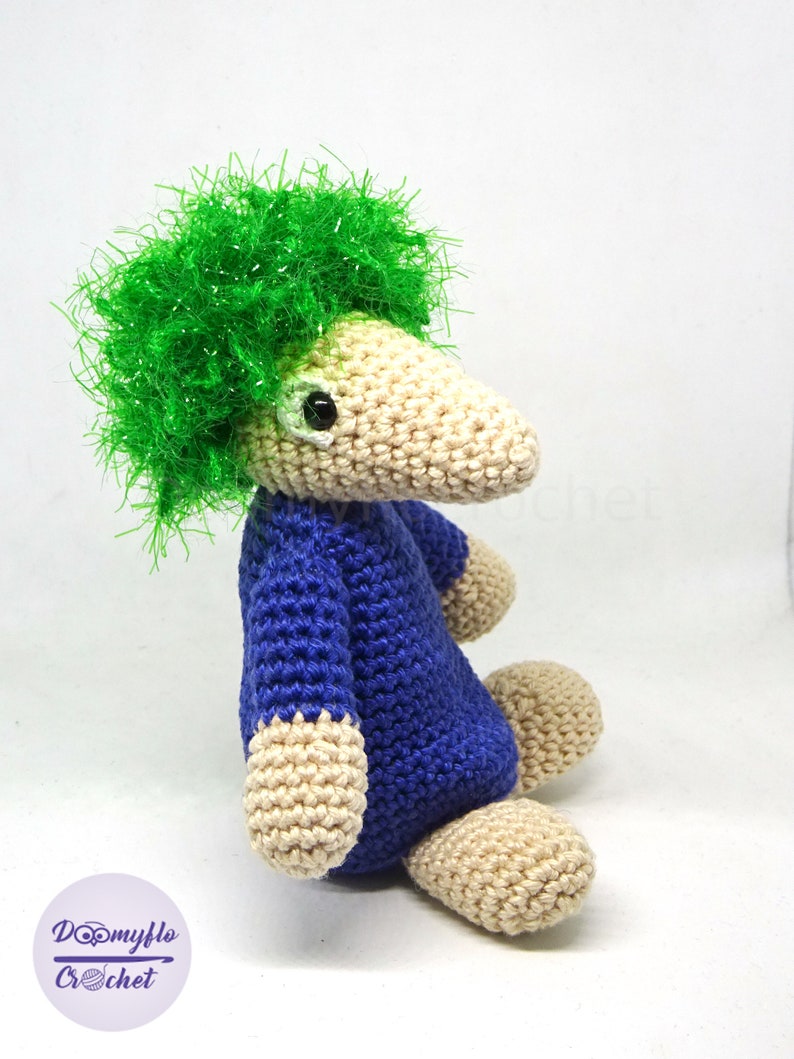 May include: A crocheted stuffed animal with a long snout, blue body, and green hair. The animal is sitting with its legs crossed.