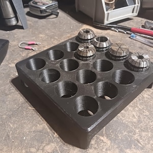 May include: A black, 3D-printed holder with 15 circular openings, designed to organise collet chucks. Several silver collet chucks are placed in the holder. A computer mouse and other tools are visible in the background.