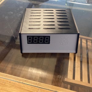 May include: A black and white electronic device with a digital display showing "8:8:8:8". The device has a rectangular shape with a vented top and a Raspberry Pi logo on the front.