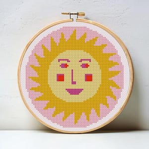 May include: A cross-stitch embroidery hoop with a smiling sun design. The sun has a yellow face with pink cheeks, a pink mouth, and pink eyes. The sun is surrounded by yellow rays and a pink background.