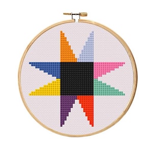 May include: A colorful cross-stitch design featuring a geometric star pattern. The star is composed of various colors, including orange, yellow, blue, pink, green, purple, and red, with a black square in the center. The design is set within a wooden embroidery hoop.