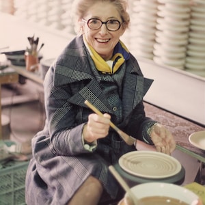 May include: A woman in glasses and a plaid coat smiles while holding a paintbrush over a pottery wheel. A yellow scarf is around her neck. Stacks of white ceramic plates are in the background. A bowl of liquid and a plate are in the foreground.