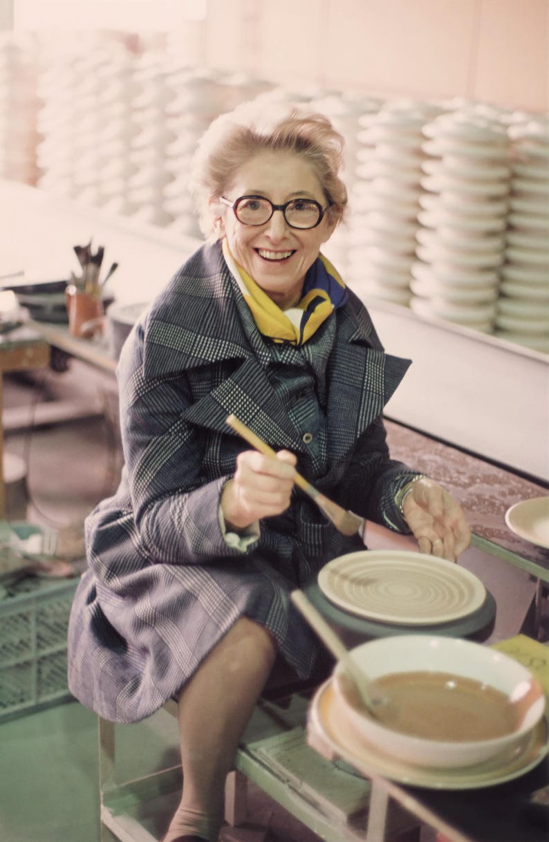 May include: A woman in glasses and a plaid coat smiles while holding a paintbrush over a pottery wheel. A yellow scarf is around her neck. Stacks of white ceramic plates are in the background. A bowl of liquid and a plate are in the foreground.