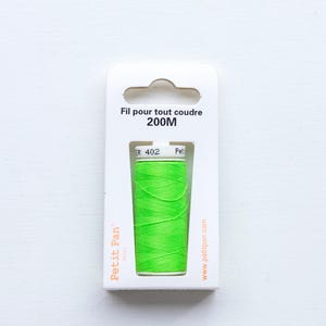 May include: A spool of bright neon green sewing thread packaged in a white cardboard holder. The packaging features the text Fil pour tout coudre 200M, the brand name Petit Pan, and the website address www.petitpan.com printed in black and orange.