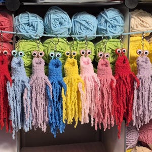 Puede incluir: Una colección de trece llaveros de pulpo tejidos a ganchillo en varios colores, incluyendo amarillo, rosa, rojo, azul, lavanda y verde. Cada pulpo presenta dos ojos móviles grandes y tentáculos largos y colgantes, sujetos a una anilla metálica.