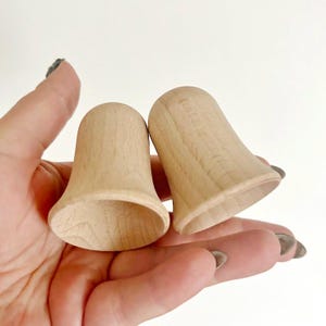 May include: Two unfinished wooden bell-shaped objects. The bells are held in a hand, showcasing their natural wood grain and smooth, rounded forms. The open bottoms and flared rims give them a classic bell shape.