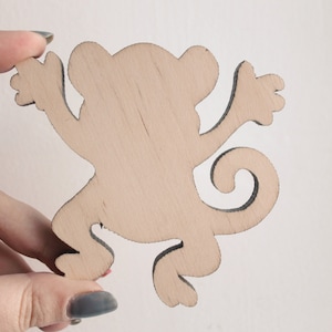 Puede incluir: Un recorte de madera de un mono en una pose juguetona. El mono es de color marrón claro y tiene un diseño simple y caprichoso.