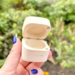 Könnte beinhalten: Kleine, offene, quadratische Holzbox mit Scharnierdeckel. Die Box ist aus hellfarbenem Holz gefertigt und hat eine glatte Oberfläche. Der Deckel ist leicht geöffnet und gibt den Blick ins Innere frei. Ein kleiner Magnet ist sichtbar.