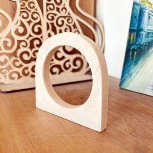 May include: A light-colored, unfinished wooden arch with a circular cutout in the center. The object has a flat base and a smooth, natural wood grain texture, standing upright as a singular, solid piece of carved wood with a rounded top edge.