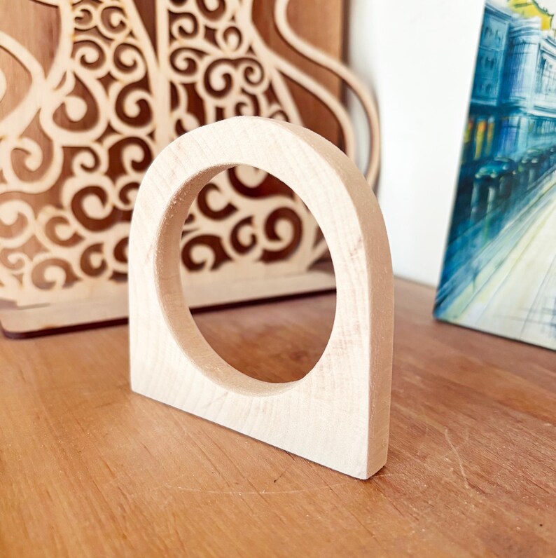 May include: A light-colored, unfinished wooden arch with a circular cutout in the center. The object has a flat base and a smooth, natural wood grain texture, standing upright as a singular, solid piece of carved wood with a rounded top edge.