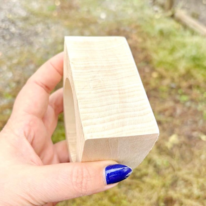 May include: A light-colored, unfinished wooden block with a smooth, rectangular exterior and a circular cutout in the center. The wood grain is visible across the surface, showing natural light beige tones and a matte, unpolished texture.