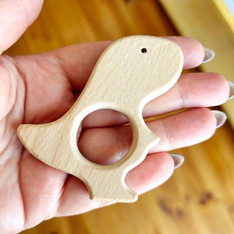 Peut inclure: Un jouet de dentition en bois clair en forme d'oiseau. Le jouet en forme d'oiseau a une grande ouverture circulaire au centre et un petit trou pr&egrave;s du sommet. Le jouet est tenu dans une main.