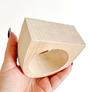 May include: A light-colored, unfinished wooden napkin ring with a square-shaped exterior and a circular interior opening. The wood grain is visible across the smooth, matte surface of the object, which features clean, sharp edges and a geometric design.