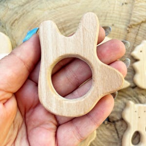 Peut inclure: Un hochet en bois clair pour b&eacute;b&eacute;, en forme de lapin stylis&eacute;. Le hochet pr&eacute;sente une grande ouverture circulaire au centre. Il est tenu dans une main, avec le grain du bois visible. D'autres jouets en bois sont en arri&egrave;re-plan.