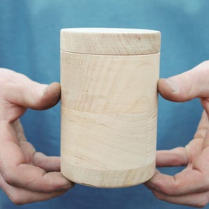 K&ouml;nnte beinhalten: Ein zylindrischer Holzbeh&auml;lter mit Deckel. Die hellfarbene Holzmaserung ist auf der Oberfl&auml;che sichtbar. Der Beh&auml;lter wird von zwei H&auml;nden vor einem blauen Hintergrund gehalten. Der Beh&auml;lter ist etwa 13 cm hoch.