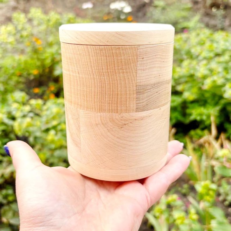 K&ouml;nnte beinhalten: Ein zylindrischer Holzbeh&auml;lter mit Deckel, gehalten in einer Hand. Der Beh&auml;lter besteht aus hellem Holz mit sichtbaren Maserungsmustern. Der Hintergrund zeigt gr&uuml;nes Laub.
