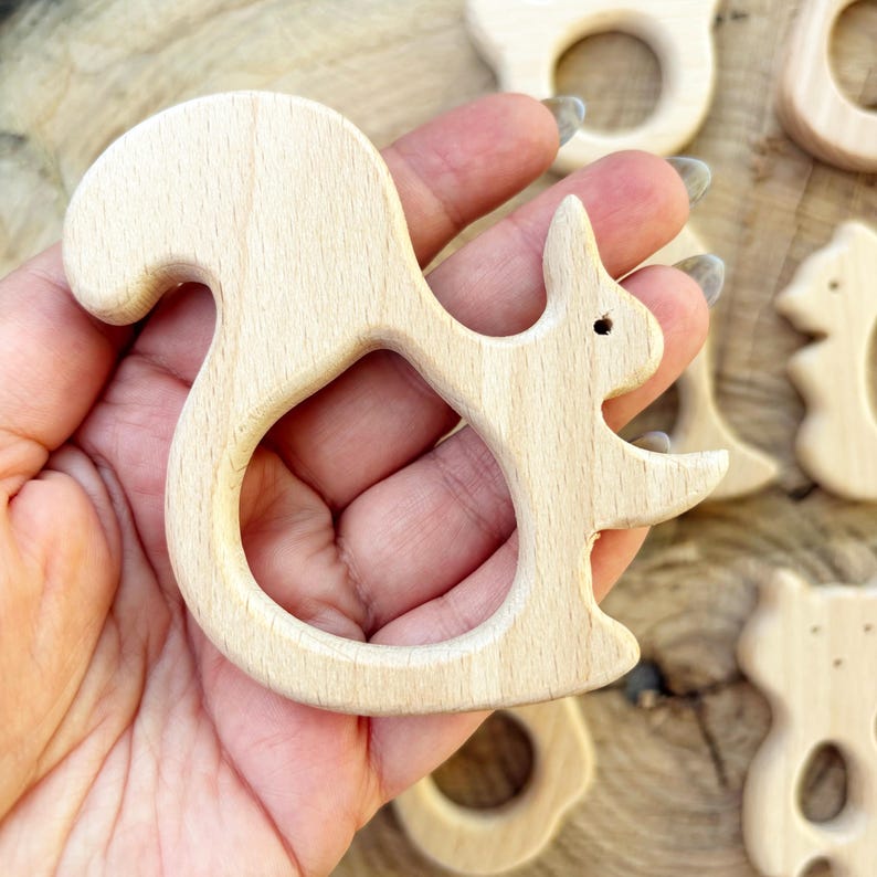 Peut inclure: Un jouet de dentition en bois clair en forme d'&eacute;cureuil. Le jouet a une finition lisse et un trou au centre pour une prise en main facile. D'autres jouets similaires sont visibles en arri&egrave;re-plan.