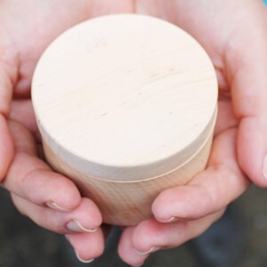 Può includere: Una piccola scatola rotonda di legno con coperchio, tenuta in mani aperte. La scatola è realizzata in legno chiaro e ha un design semplice e naturale. La scatola misura circa 5 cm di diametro e 4 cm di altezza.
