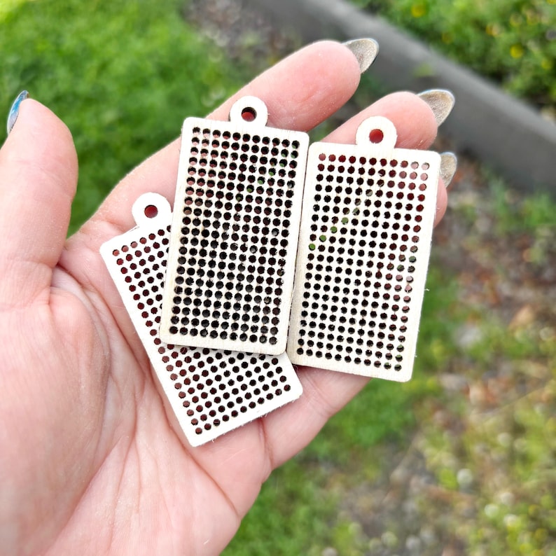 Peut inclure: Trois pendentifs rectangulaires en bois clair, chacun pr&eacute;sentant une grille uniforme de petites d&eacute;coupes circulaires et un trou unique en haut pour la suspension. Les pendentifs ont une surface plate et lisse avec une finition en bois beige.