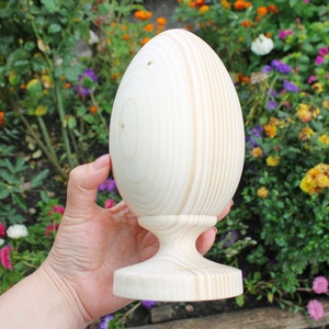 May include: A large, unfinished, wooden egg-shaped ornament with a round base. The ornament is made of light-colored wood and has a smooth, polished finish.