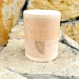 K&ouml;nnte beinhalten: Ein zylindrischer Holzbeh&auml;lter mit Deckel. Der Beh&auml;lter besteht aus hellfarbenem Holz mit sichtbaren Maserungsmustern. Der Deckel hat eine einfarbige wei&szlig;e Farbe. Der Beh&auml;lter steht auf einer Steinoberfl&auml;che.